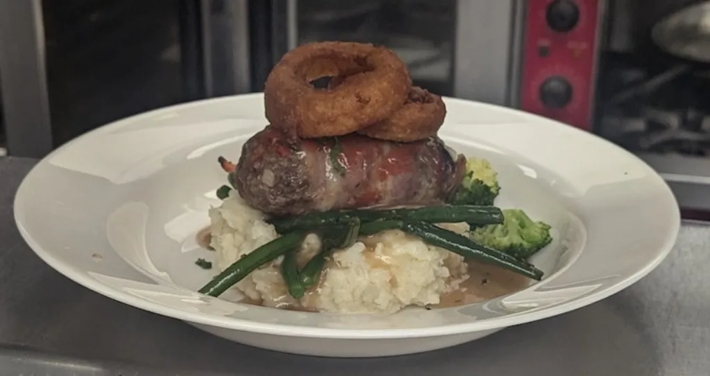 Hearty bacon-wrapped meatloaf with mashed potatoes and onion rings.